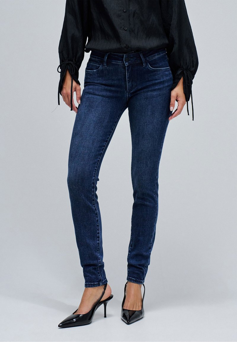 Kobieta ubrana w ciemnoniebieskie obcisłe dżinsy, czarne szpilki z ostrym noskiem oraz czarną bluzkę z długim rękawem i wiązanymi mankietami.