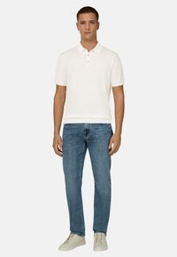 Homme debout face à la caméra, portant un polo blanc à manches courtes, un jean bleu et des baskets beige, sur un fond uni.