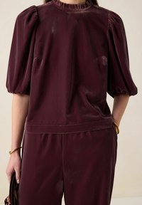 Haut en velours bordeaux avec manches bouffantes et encolure froncée, assorti à un pantalon coordonné. Texture lisse et détail subtil de brillance.