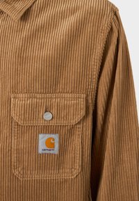 Brązowa koszula z korduroyu z kieszenią zapinaną na guzik i naszywką z logo Carhartt. Charakteryzuje się pionowym żebrowaniem oraz klasycznym krojem kołnierza.