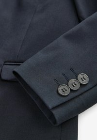 Manche de blazer bleu marine foncé avec trois boutons noirs cousus près du poignet et une petite poche visible en arrière-plan.