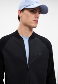 Svart zip-up jakke med teksturerte svarte ermer, brukt over en lyseblå t-skjorte. En blå caps fullfører antrekket, med en logo på forsiden.