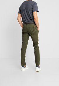 Olivově zelené slim-fit chinos s rovnými nohavicemi, vyrobené z bavlněného twillu, se zadními kapsami a černým páskem, v kombinaci se šedou košilí.
