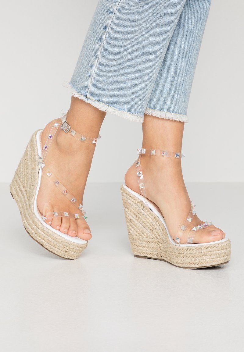 Sandales à talons transparents en forme de coin avec des accents géométriques argentés, un talon en jute tissé et une semelle plateforme blanche ; les brides de cheville sont ornées de clous.