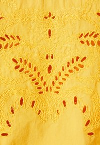 Gult tyg med intrikata blomstermönster och utskurna detaljer i en kontrasterande orange, vilket skapar en texturerad och detaljerad design.