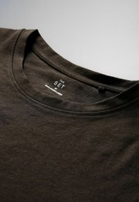 Camiseta marrón oscuro con cuello redondo, con cuello cosido y etiqueta impresa "THE SET M MADE IN BANGLADESH" dentro del escote.