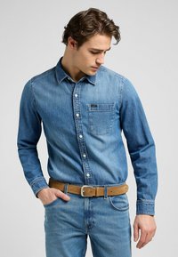 Denim overhemd in lichtblauw met een knoopsluiting aan de voorkant, een zak op de borst en lange mouwen, gecombineerd met een bruine riem en bijpassende jeans.