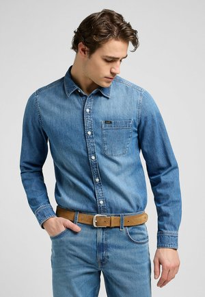Chemise en denim bleu clair avec une fermeture à boutons, une poche sur la poitrine et des manches longues, associée à une ceinture marron et un jean assorti.