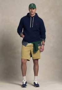 Tengerész kék kapucnis pulóver fehér húzózsinórral, világos kockás ing rétegként, khaki rövidnadrág, zöld pulóver a derékon megkötve, és kék sportcipő fehér részletekkel.