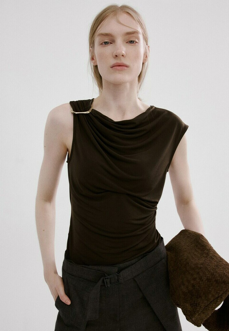 Plava žena u crnoj top bez rukava s drapiranim detaljem i zlatnim kopčama, tamnim hlačama s visokim strukom, drži presavijenu smeđu tkaninu teksture.