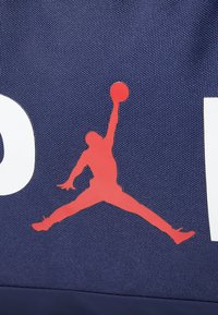 Bolsa azul marino con el logo rojo de Jumpman, que presenta una tela texturizada y letras blancas audaces. El diseño destaca una estética deportiva y elementos de marca.