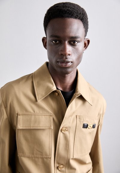Tan work jacket with a structured collar, two front pockets, gold buttons, and a decorative badge detail. Smooth fabric with a matte finish.