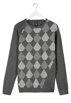 Mand i grå blazer og mønstret sweater, der holder en sort og brun læder-accent taske, iført sorte bukser og beige sko.