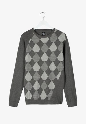 Homme en blazer gris et pull à motifs tenant un sac noir et marron avec des accents en cuir, portant un pantalon noir et des chaussures beiges.