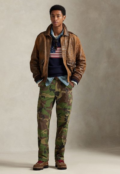 Bruine leren bomberjack over een marineblauwe trui met een Amerikaanse vlag, denim overhemd, camo cargobroek en stoere veterlaarzen.