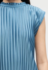 Κοντινή λήψη πλισέ μπλε αμάνικου τοπ με βολάν στο ντεκολτέ που φοράει άτομο με μακριά σκουρόχρωμα μαλλιά.