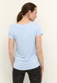 Jasnoniebieska koszulka z krótkim rękawem i szerokim dekoltem, o gładkiej fakturze i delikatnym kroju. Noszona z dopasowanymi czarnymi spodniami.