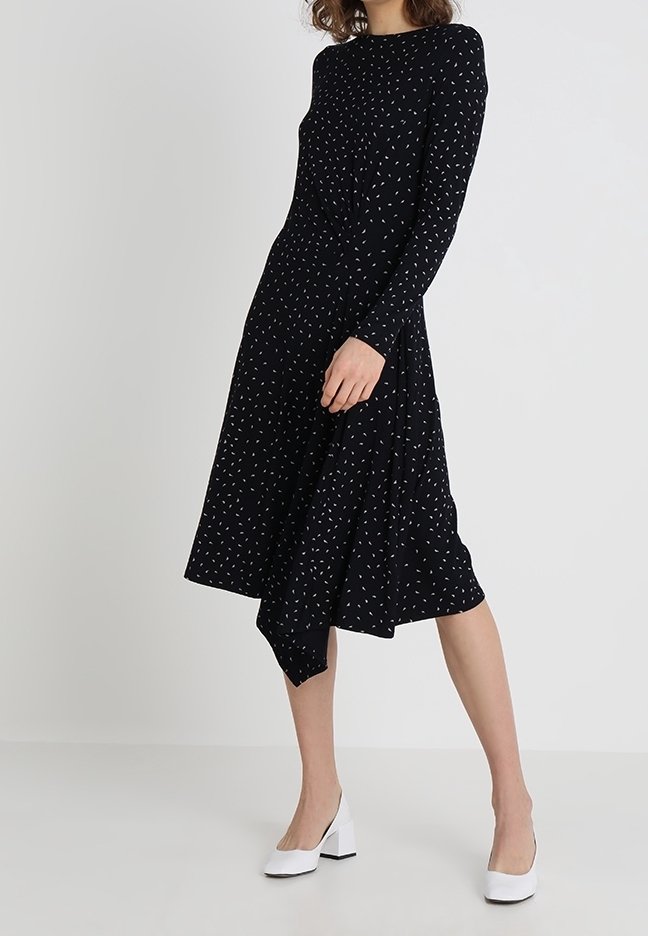 Femme portant une robe midi noire à manches longues avec de petits motifs blancs et des chaussures blanches à talons blocs, debout devant un fond uni.