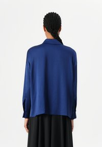 Chemise bleue surdimensionnée avec une texture lisse, dotée d'un col et de manches longues. Le dos présente un ourlet incurvé et des fentes sur les côtés.