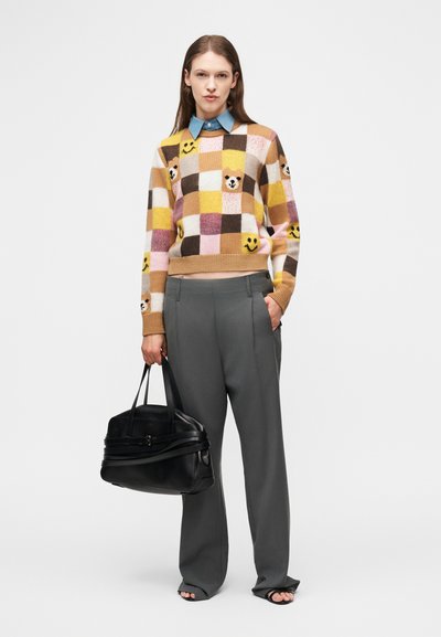 Femme portant un pull à carreaux avec des visages souriants et des ours, un pantalon gris à jambes larges, des sandales noires, tenant un grand sac à main noir.