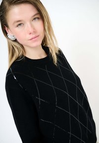 Giovane donna con capelli biondi bagnati che indossa orecchini argentati vistosi e un maglione nero con un sottile motivo a griglia diagonale su sfondo chiaro.