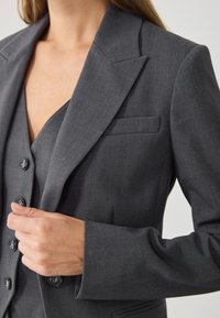 Γκρι προσαρμοσμένο σακάκι με γωνιακά πέτα, λεία υφή, μονή τσέπη στο στήθος και κλείσιμο με πέντε κουμπιά. Το ύφασμα φαίνεται απαλό και διαρθρωμένο.
