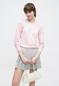 Ροζ ανοιχτόχρωμο τοπ με μακριά μανίκια σε συνδυασμό με γκρι πλισέ μίνι φούστα. Κρατά μια μικρή, λευκή τσάντα με καμπυλωτές λαβές.