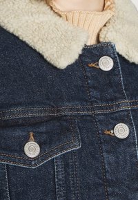 Denim jacket with beige sherpa collar, visible buttons embossed with "Tommy Jeans New York," and beige knit sweater underneath.