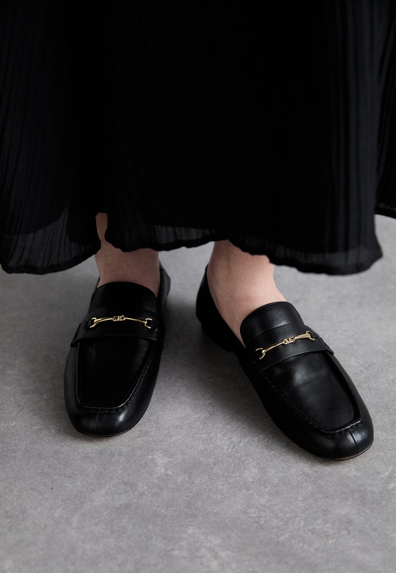 Zwarte leren loafers met een gestructureerde afwerking, voorzien van een gouden accent over de wreef en een vierkante neusvorm.