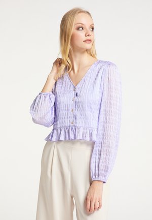Femme aux cheveux blonds portant un chemisier texturé violet clair à manches longues avec des boutons et un pantalon beige taille haute, regardant sur le côté.