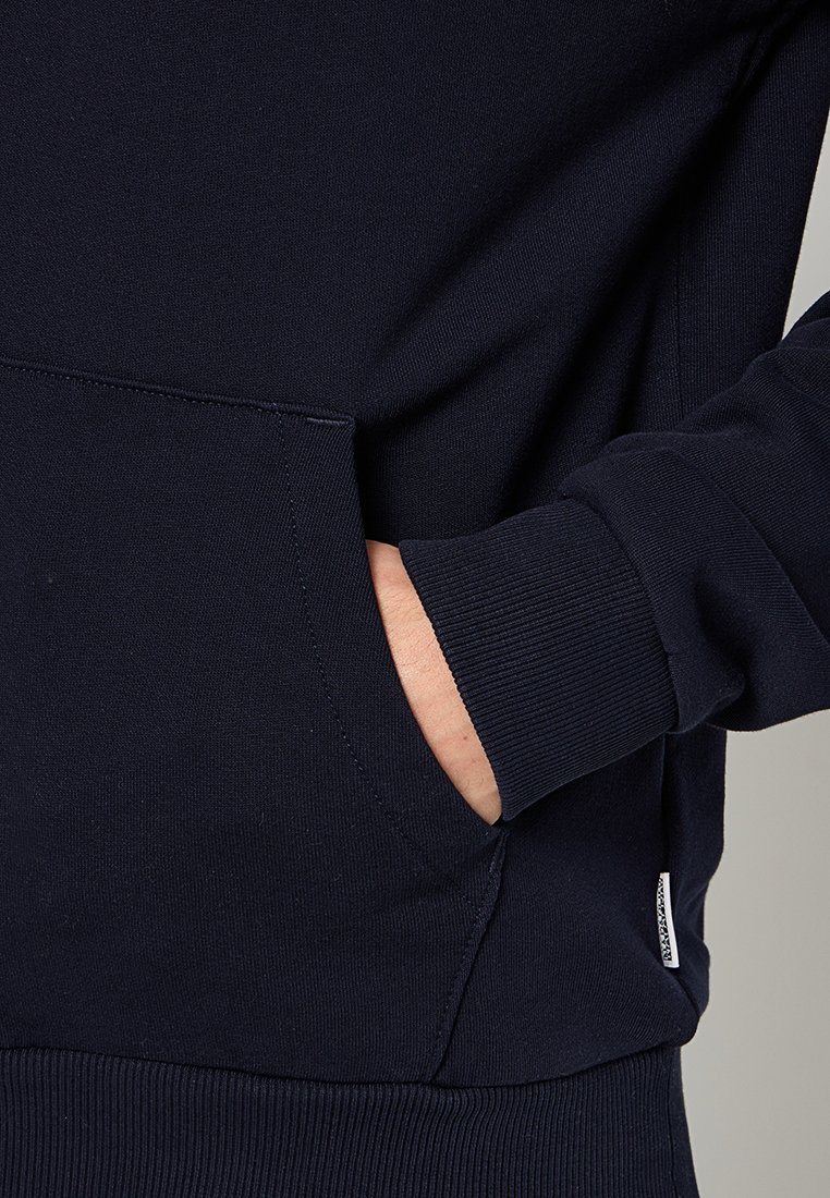 Marineblaue Sweatshirt aus strukturiertem Stoff, mit einer Fronttasche und gerippten Bündchen, sowie einem kleinen Etikett am Saum.
