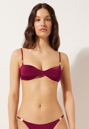 Mujer con cabello largo y ondulado, vistiendo un bikini magenta oscuro con detalles dorados, de pie contra un fondo liso color marfil.