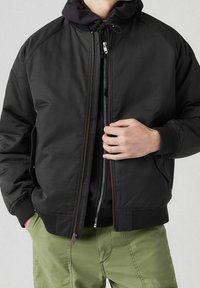 Svart bomberjacka med dragkedja fram, ribbade muddar och sidofickor. Bärs över en svart huvtröja, i kombination med olivgröna shorts.