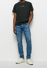 Camiseta negra de algodón con logo blanco, combinada con jeans de mezclilla azul claro desgarrados y zapatillas blancas hueso, con un corte clásico y un estilo casual.