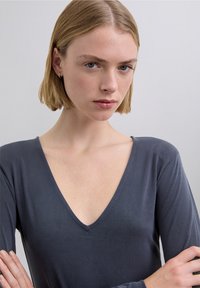 Top gris de manga larga con escote en V profundo, textura suave y diseño sencillo. La modelo tiene los brazos cruzados, mostrando el ajuste de la prenda.