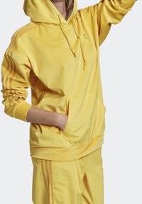 Gul huvtröja i ribbad tyg. Har en framficka, dragsko och kontrasterande ränder längs ärmarna.