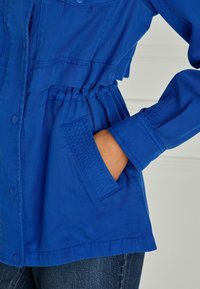 Hand in hip pocket of blue jacket with cinched waist and buttoned front, paired with dark denim jeans on a light background.