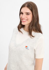 Jongeman met schouderlang bruin haar, gekleed in een witte shirt met korte mouwen en een fuzzy mouwloze vest met een kleurrijke logo-patch.