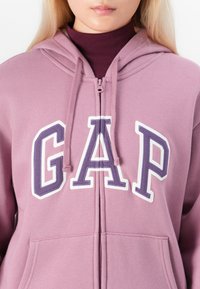Roze ritsjack met een groot "GAP"-logo in wit en donkerpaars, met trekkoorden en voorkwasten. Zachte stofstructuur.