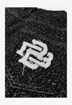 Zwarte textuurstof met een witte geborduurde logodetail. Het logo heeft rafelige randen. Zichtbare stiksels op het kledingstuk.