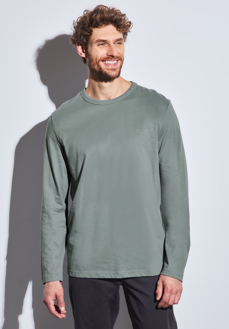 Camisa de manga larga de color verde claro hecha de tela suave, con cuello redondo y un sutil detalle de logo. Combinada con pantalones oscuros.