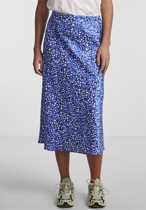 Kvinde iført en blå midinederdel med hvide og sorte pletter, hvid t-shirt og beige sportssko med grønne accenter på en ensfarvet baggrund.