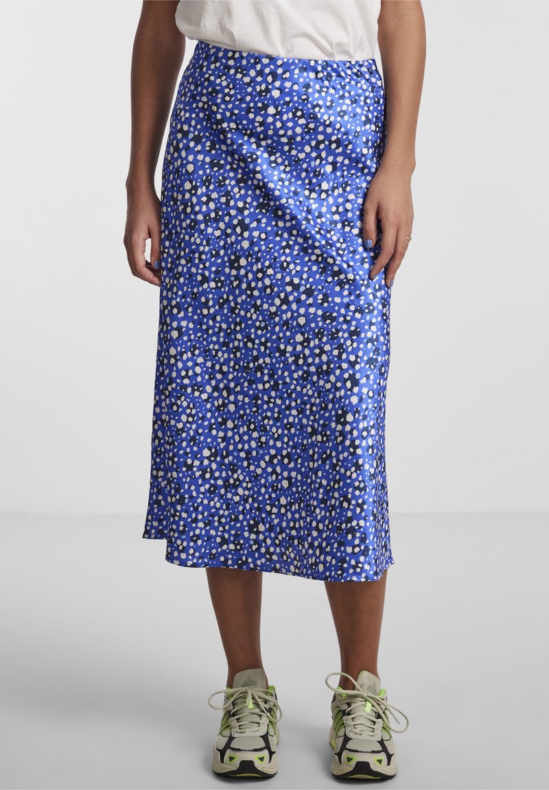 Femme portant une jupe midi bleue avec des taches blanches et noires, un t-shirt blanc, et des chaussures de sport beiges avec des accents verts sur un fond uni.
