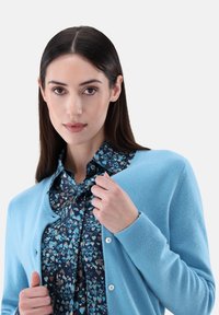 Hellblauer Cardigan aus weichem Material mit Rundhalsausschnitt und Knopfverschluss, getragen über einer gemusterten Bluse in Blau und Marineblau.
