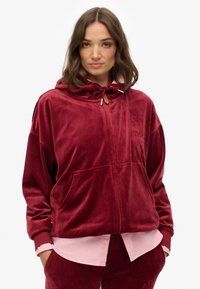 Haut à capuche en velours zippé rouge profond avec un cordon de serrage, poches avant et détail de logo brodé. Porté par-dessus une chemise rose clair.