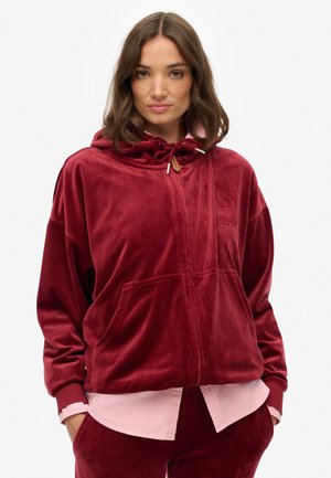 Velvet zip-up hoodie i dyb rød med snørehætte, forlommer og broderet logo-detalje. Båret over en lyserød skjorte.