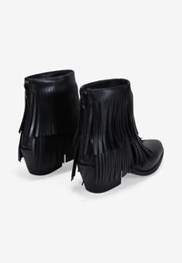 Botas de tobillo de cuero negro con largos flecos, cremalleras laterales y un tacón bajo apilado, con una textura suave y un diseño moderno.