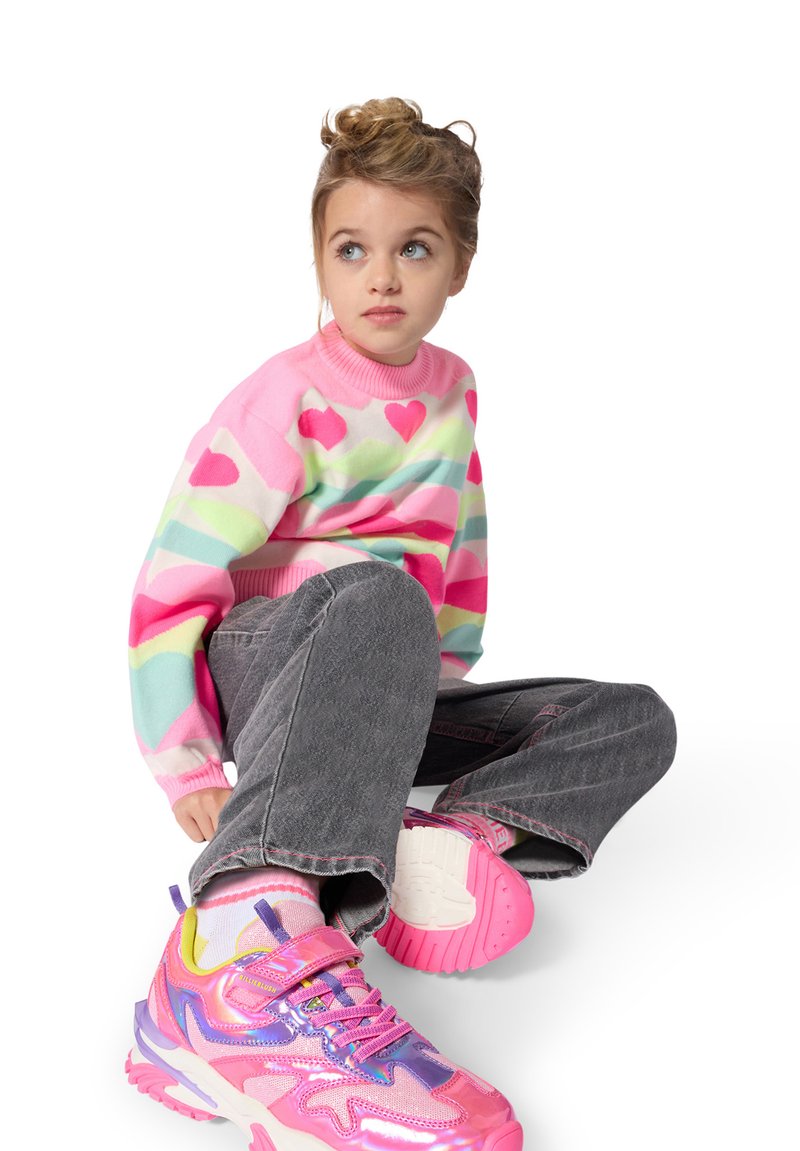 Jeune fille aux cheveux blonds assise, portant un pull coloré à motif de cœur, un pantalon gris et des baskets roses et violettes éclatantes.