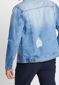Man in een lichtblauwe denimjas met een versleten patch op de onderrug, gecombineerd met donkere gestreepte pantalons.
