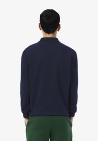 Polo shirt à manches longues bleu marine avec col et poignets élastiques, associé à un pantalon vert, vu de dos. Texture douce, coupe décontractée.